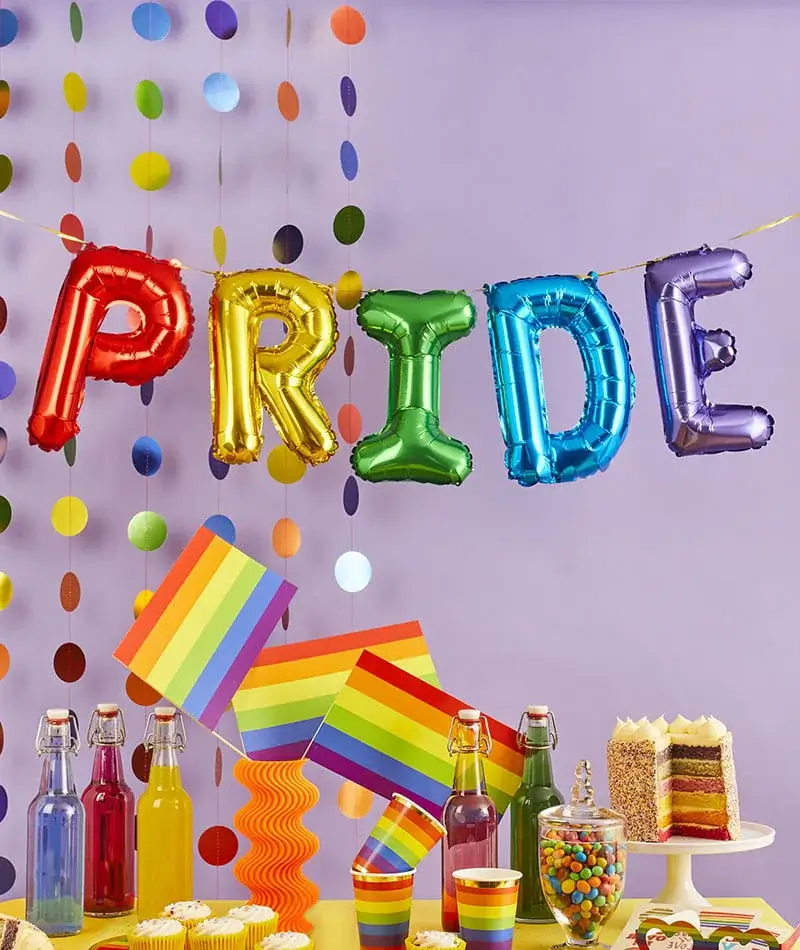 Rainbow Pride party setup with colourful PRIDE balloon letters, rainbow flags, drinks, cupcakes, and a layered cake on a decorated table